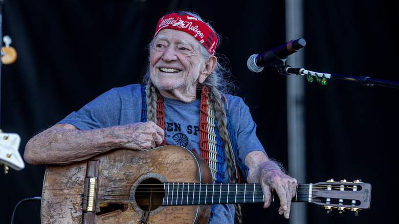 Willie Nelson smiles on stage, holding a guitar in front of a microphone.