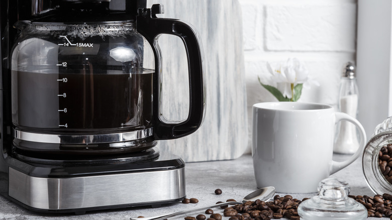 A drip coffee maker full of coffee with a white mug beside it