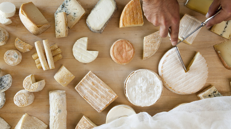 Various types of cheeses on a wooden surface while a man cuts into a block of brie