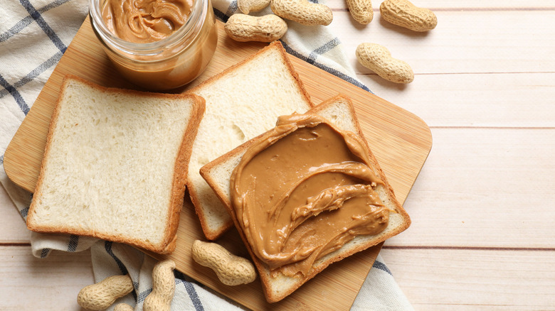 Peanut butter spread on white sandwich bread alongside whole peanuts and jar of peanut butter