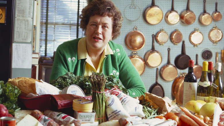 Julia Child in her kitchen by a table covered with fresh produce.