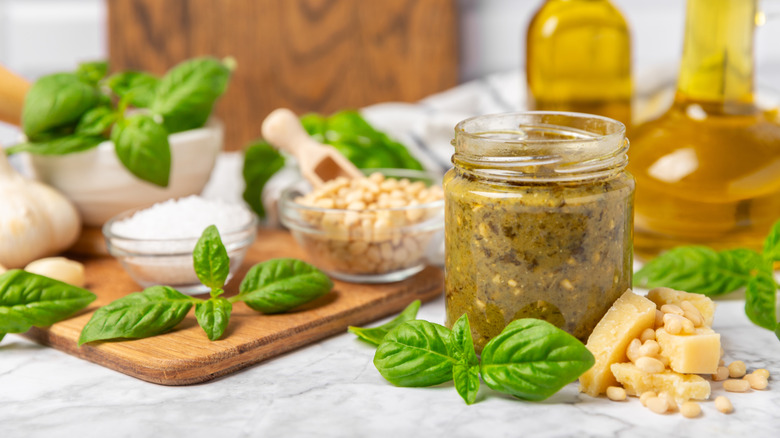 A small jar of pesto surrounded pine nuts, garlic, olive oil, Parmesan cheese, and basil