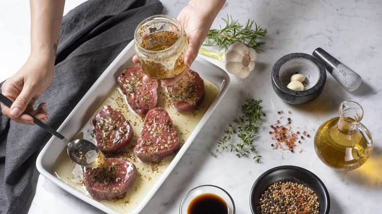 Chef spoons marinade over filets in a ceramic baking dish, with olive oil and herbs nearby