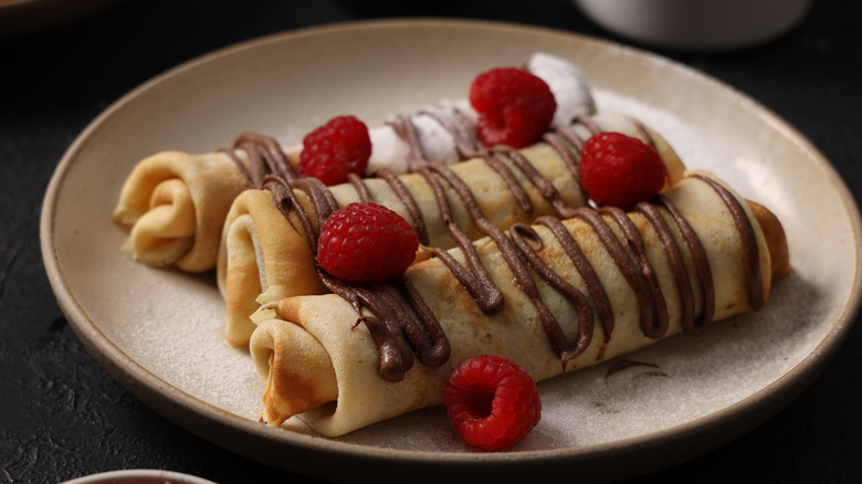 Three rolled up crepes garnished with chocolate drizzle and raspberries