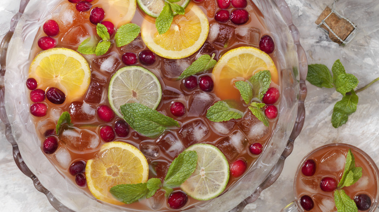 Glass punch bowl full of rum punch, ice, mint leaves, lime and orange slices as well as cherries.