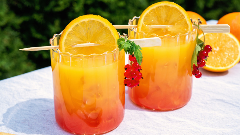 Rum punch ombré from red to yellow with an orange slice as garnish in clear glasses on a white surface and green background.