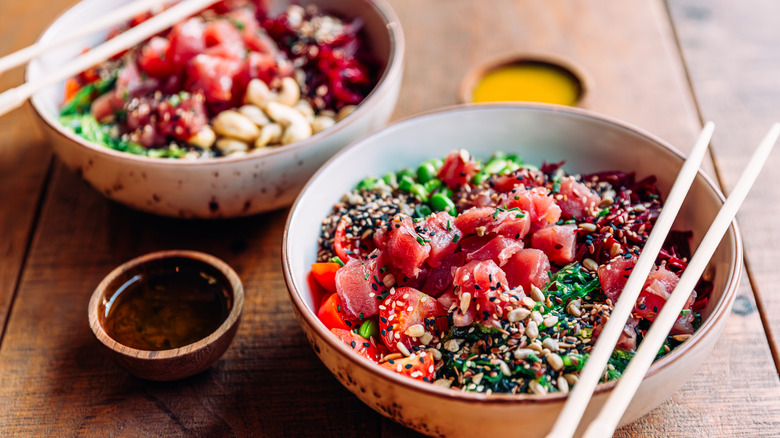 Two bowls of poke next to one another with chopsticks resting on them on a wooden surface