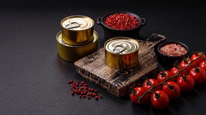 Gold cans of food, tomatoes, peppercorns, and a wood board on a dark surface