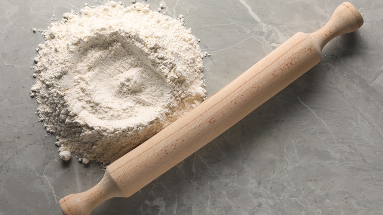 A wooden rolling pin next to a mound of flour on a grey surface