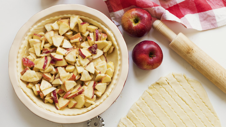 A pie dish lined with pastry and chopped apples with the pie lid and whole apples to one side