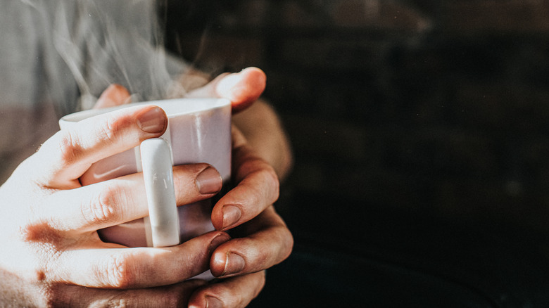 person holding warm steaming white mug in hands on black background