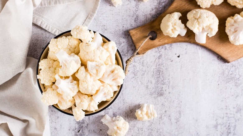 A bowl of cauliflower next to a cutting board with cauliflower on it