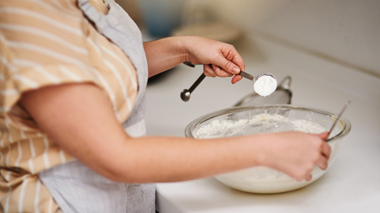 A person holding a measuring spoon full of flour above a mixing bowl