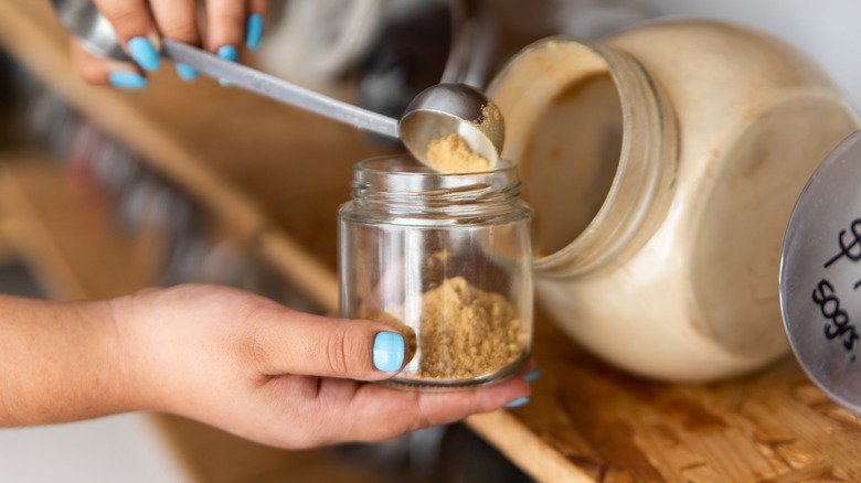 A person measuring brown sugar into a glass jar using a metal measuring spoon