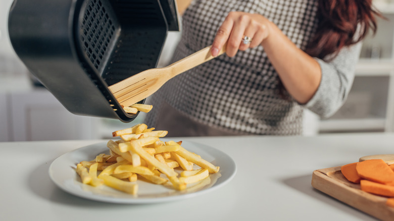 A woman pours french fries from an air fryer basket onto a plate