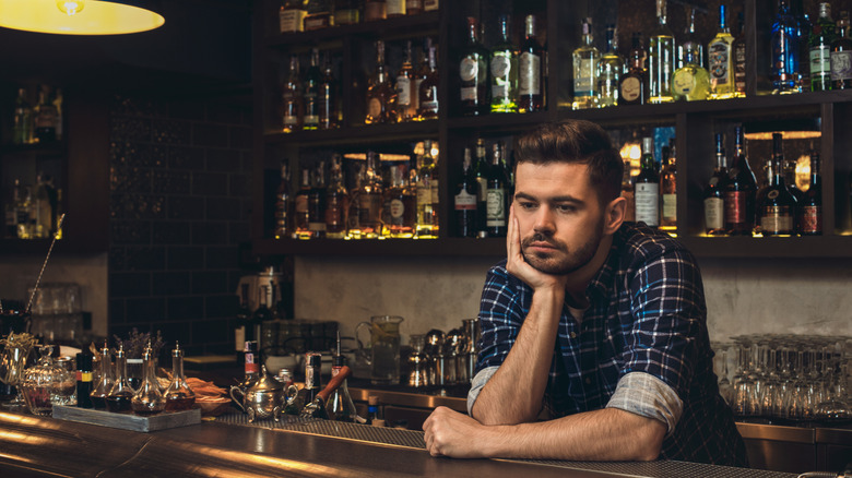 Tired barman stands behind bar resting his head on hand