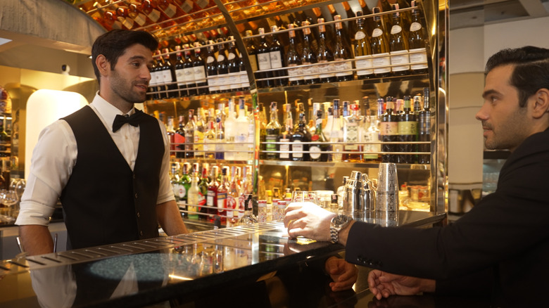 Formal bartender speaking with client next to shelves of bottles