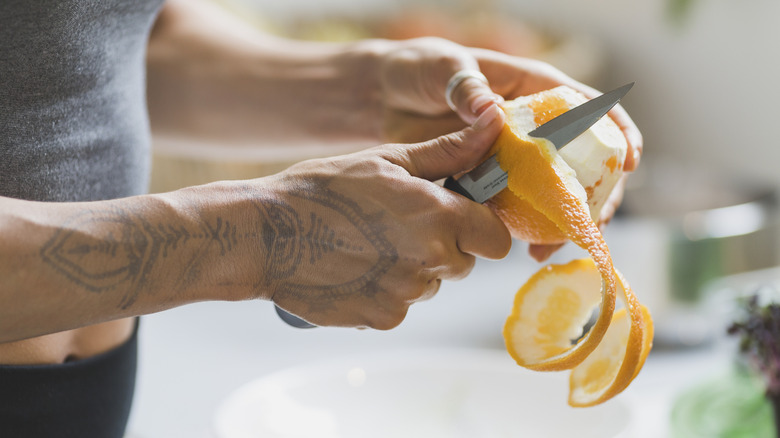 A person with tattoos peeling an orange with a knife