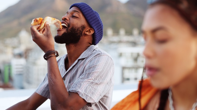 A mean eats a slice of pizza next to a woman