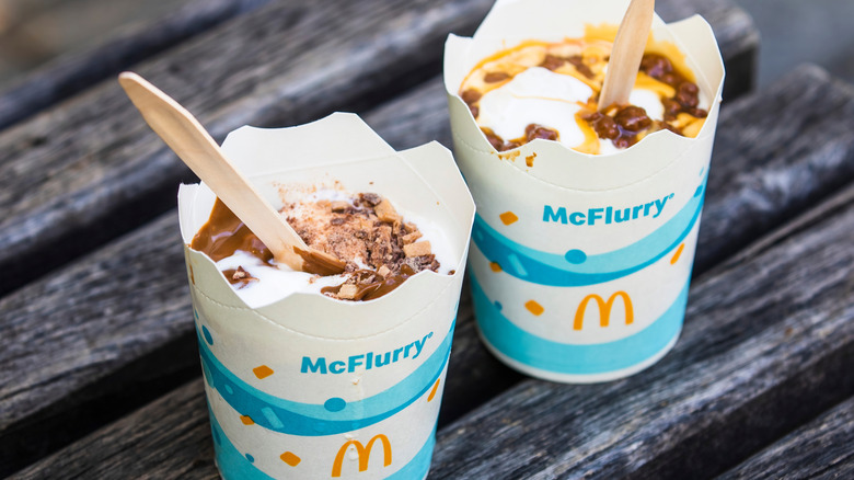 Two McFlurry cups one with chocolate sauce and one with caramel sauce on the ice cream, each with a wooden spoon.