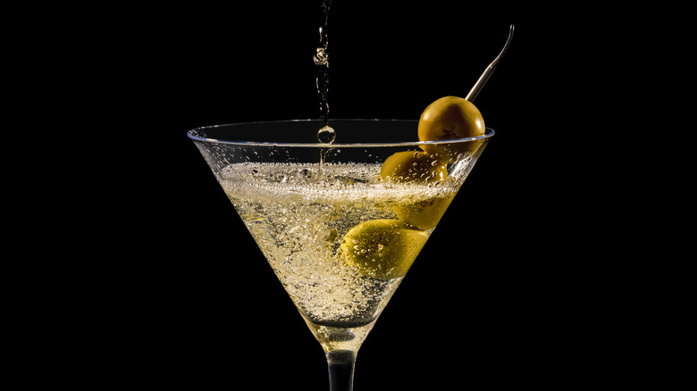 A martini being poured from off-camera into a martini glass, with three olives on a stick.