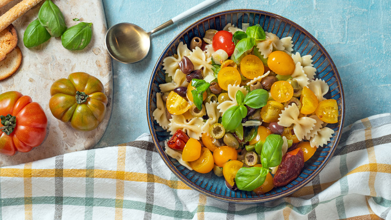 A bowl of pasta salad sitting next to herbs and tomatos