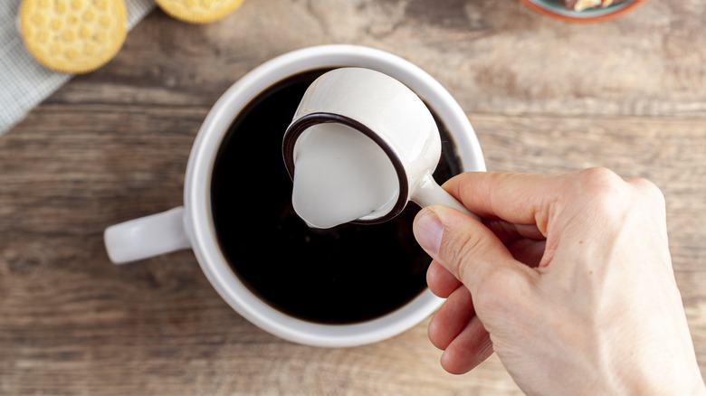 Hand pouring creamer into cup of black coffee