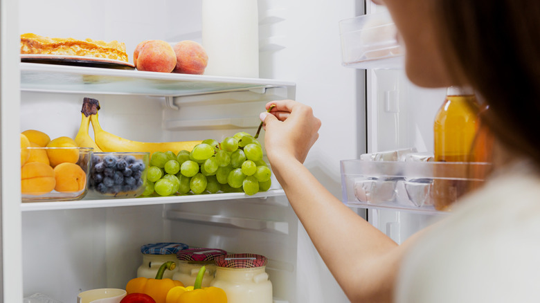 A person standing in front of an open fridge reaching for a bunch of green grapes