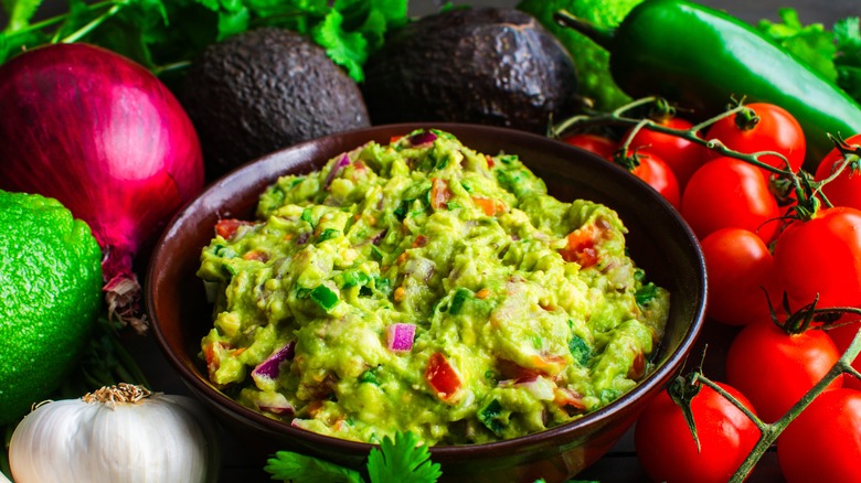 Bowl of guacamole surrounded by cherry tomatoes, avocados, lime, red onion, garlic and chilli peppers.