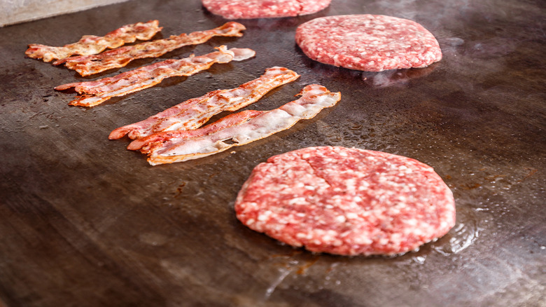 Five strips of bacon and four hamburger patties frying on a grill