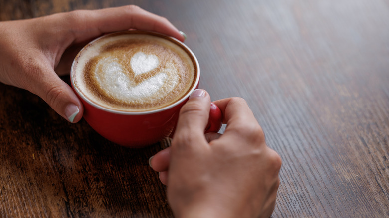 A close-up of someone's hands holding a cup of coffee with foam on top in a heart-shaped design