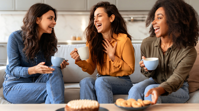 Three women sitting on a couch laughing and drinking coffee