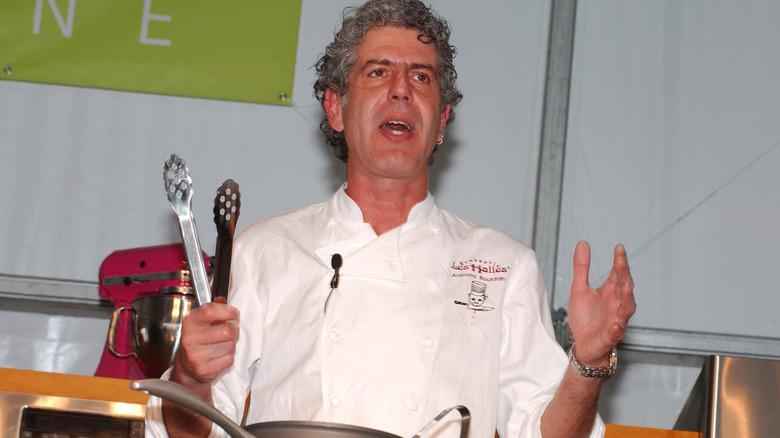 Anthony Bourdain in a chef's jacket, holding tongs.