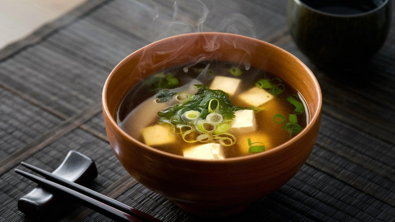 A bowl of miso soup in a brown bowl alongside black chopsticks