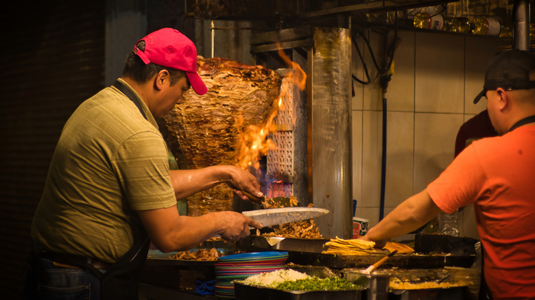 Two chefs work slicing Al Pastor meat off a spit