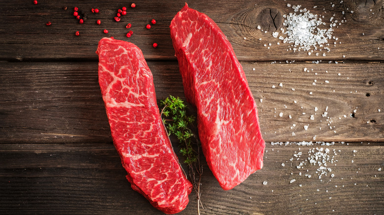 two cuts of raw denver steak on a wooden background with salt, herbs, and spices