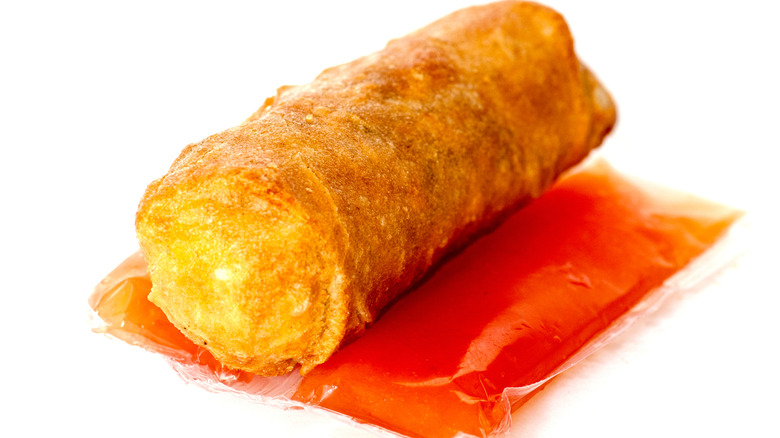 A crispy egg roll sitting on two packets of duck sauce against a white background