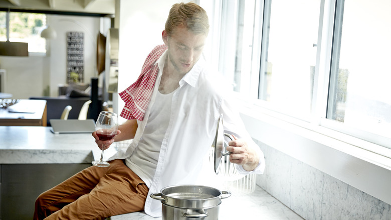 Man sitting on his counter top holding a glass of red wine and checking on his pot on the stove top