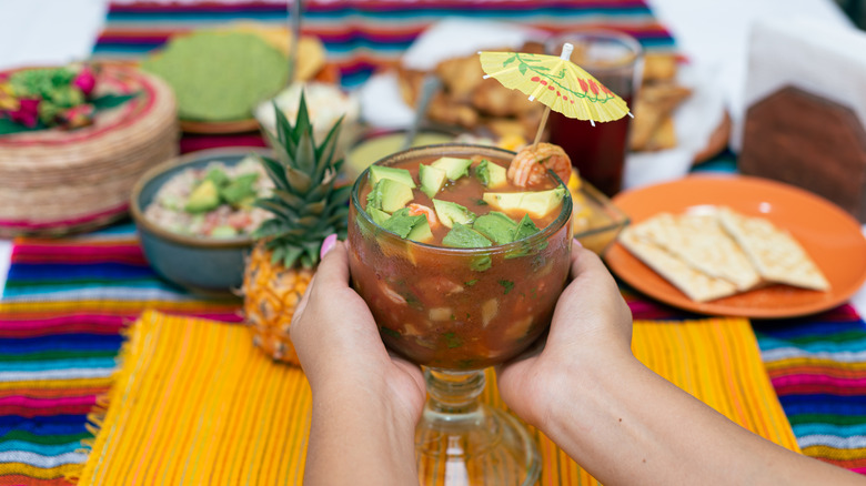 Hands holding a goblet of Mexican shrimp cocktail with umbrella garnish