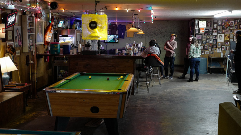 people lounge and chat in a dive bar setting featuring small bar and pool table