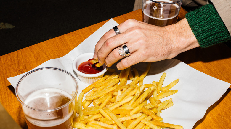 hands dipping french fries in ketchup alongside two pints of beer at a bar