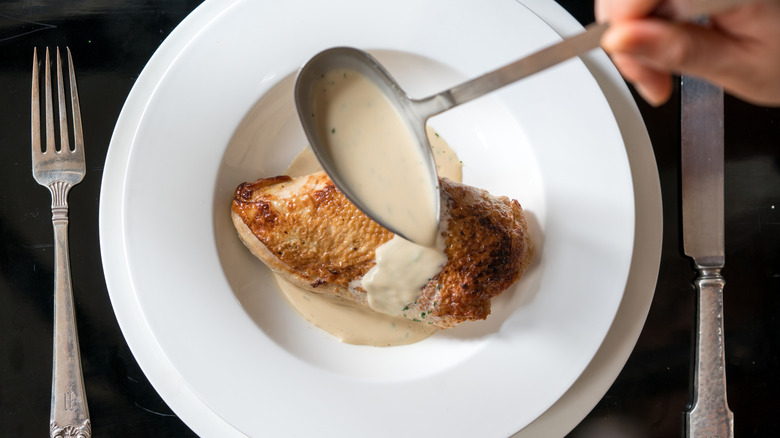 Velouté sauce being spooned over cooked chicken breast on a plate