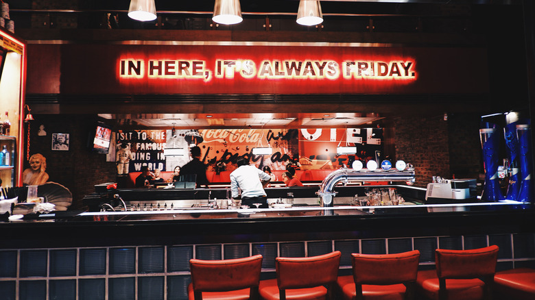 The bar inside a TGI Fridays with a red neon sign, servers making drinks, and red bar stools