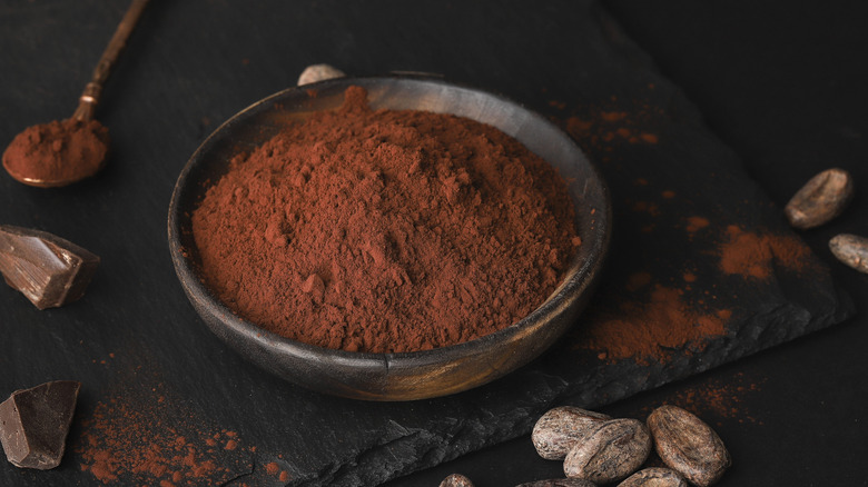 Cocoa powder in a bowl with cacao beans and broken chocolate pieces around