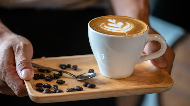 A person holding a wooden tray with a cup of coffee, stirring spoon, and coffee beans on it