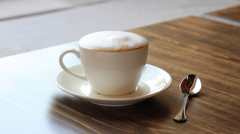 A cappuccino in a white mug with a silver spoon beside it on a wooden table