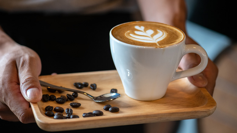 A server carrying a latte with foam in it on a wooden tray