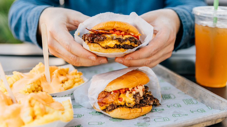 A person holding a bacon cheeseburger, with cheese fries, and a beverage nearby