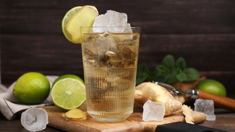 A glass of ginger ale with ice and a lime garnish surrounded by clumps of fresh ginger and limes