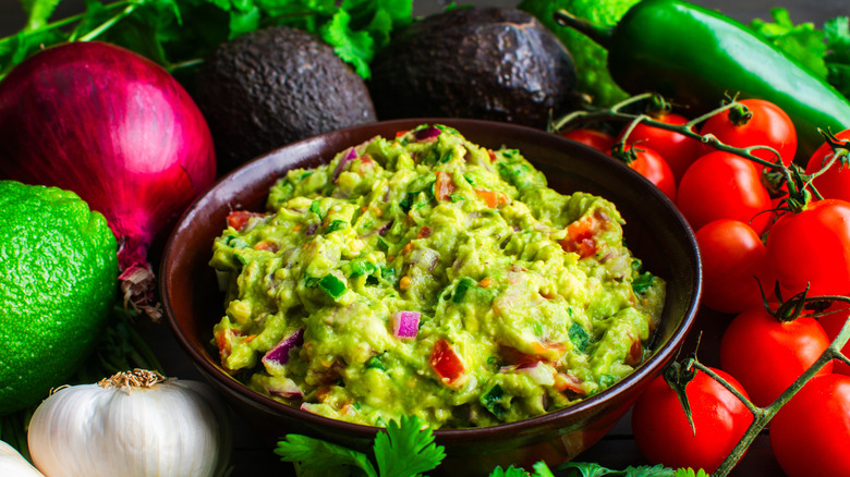 Bowl of guacamole surrounded by fruits and vegetables such as red onion, tomatoes, avocados, a jalapeno, garlic, and a lime